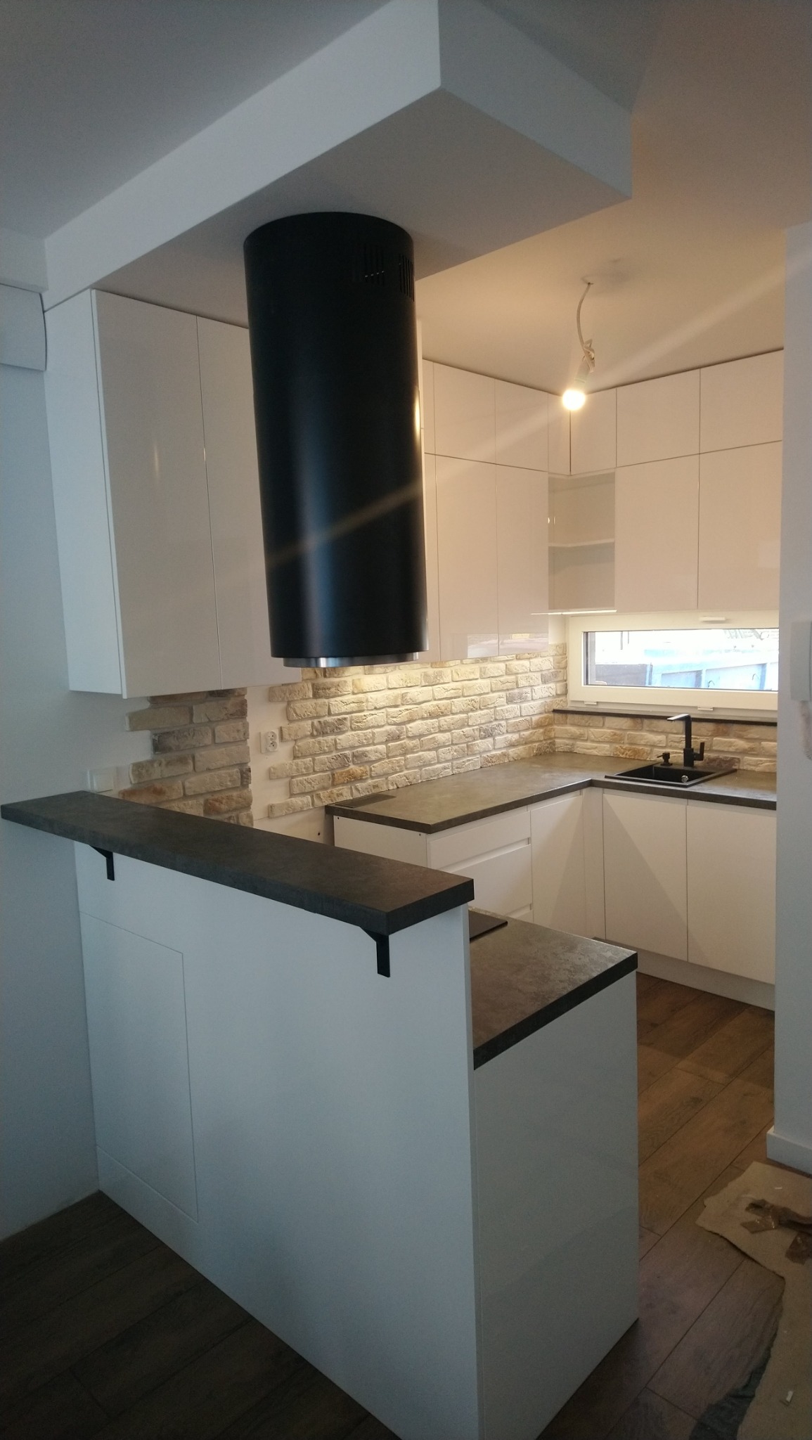 Nowoczesna, biała kuchnia z czarnym okrągłym okapem, ceglaną ścianą nad blatem i barkiem z ciemnym blatem.