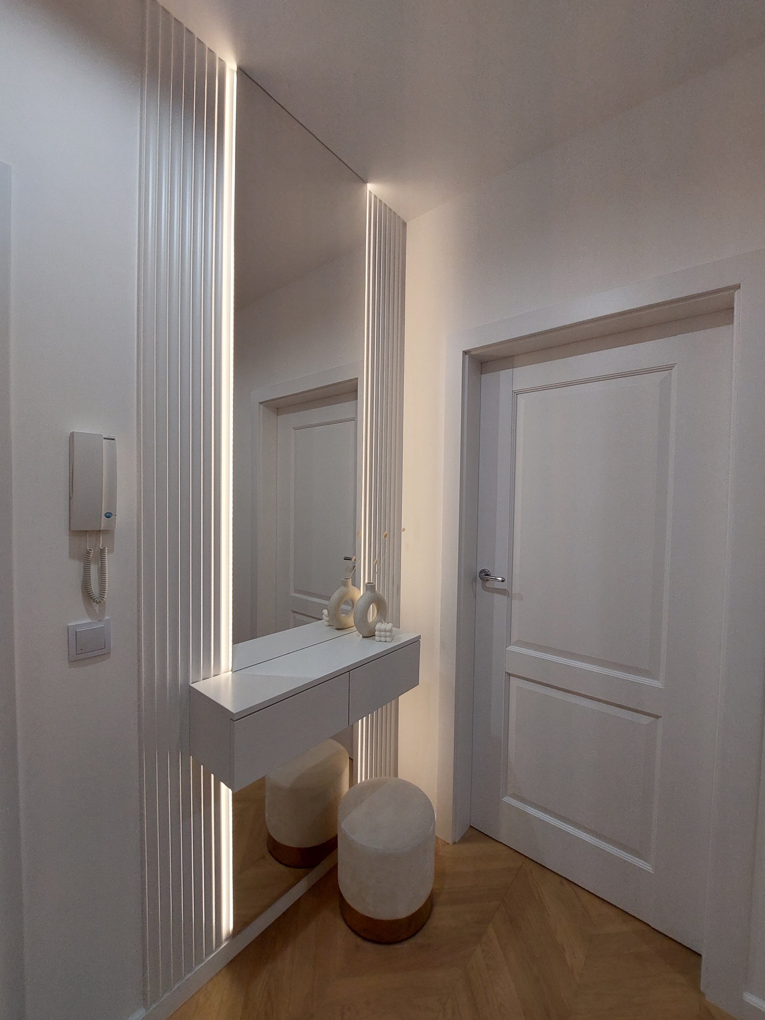 Nowoczesna aranżacja przedpokoju z białą toaletką wiszącą, lustrem z podświetleniem LED, pufem i parkietem w jodełkę.