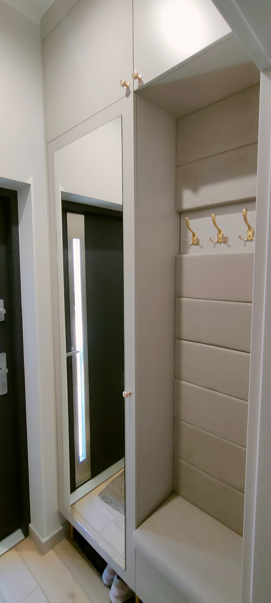 Jasnoszara szafa wnękowa z lustrem, wewnątrz wieszaki i tapicerowane panele ścienne, widoczne buty w odbiciu lustrzanym.