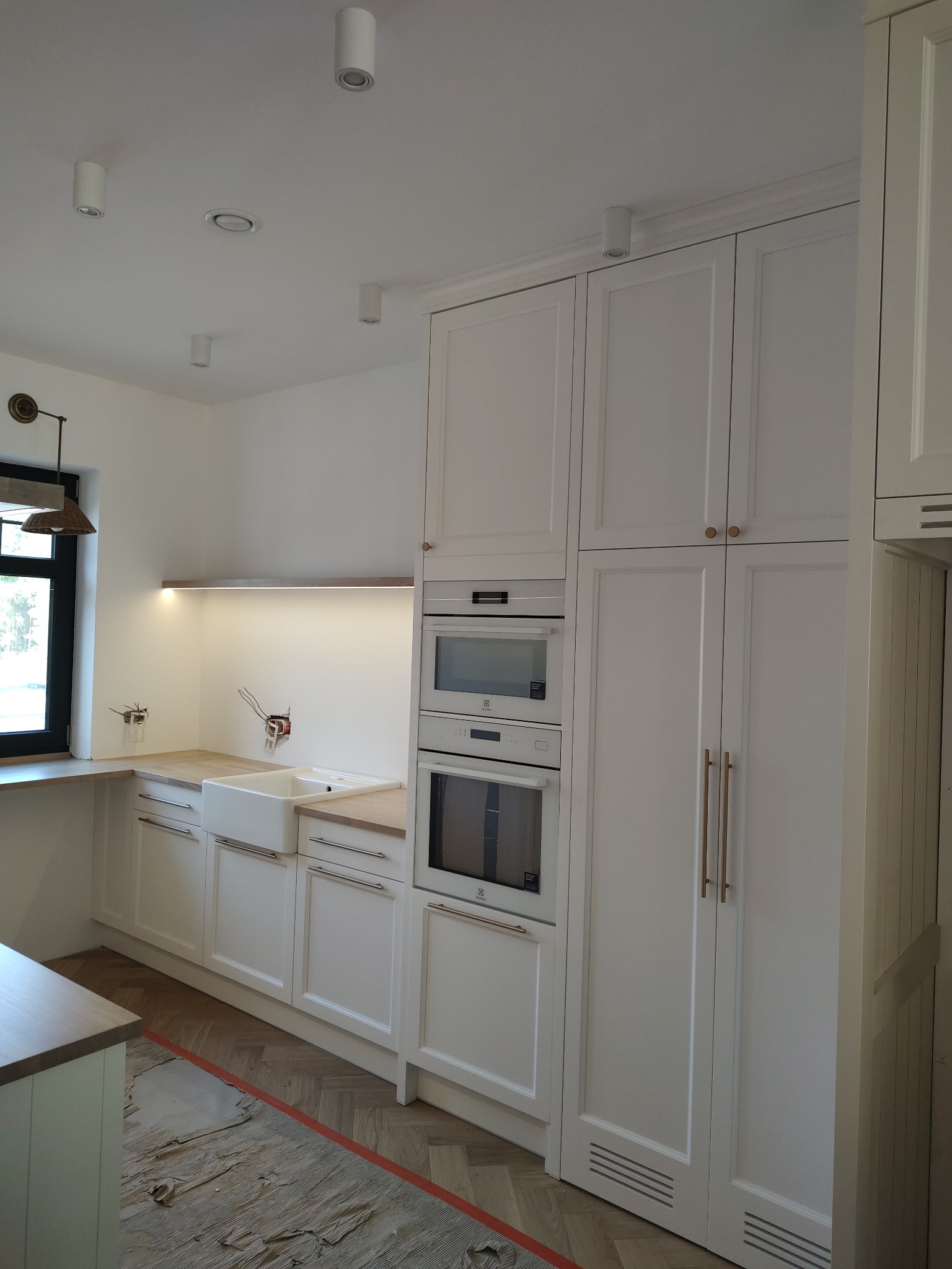 Wnętrze kuchni w trakcie remontu, z białymi szafkami, drewnianym blatem, białym zlewem, wbudowanymi sprzętami AGD marki Electrolux i częściowo odsłoniętą podłogą z parkietu ułożonego w jodełkę.