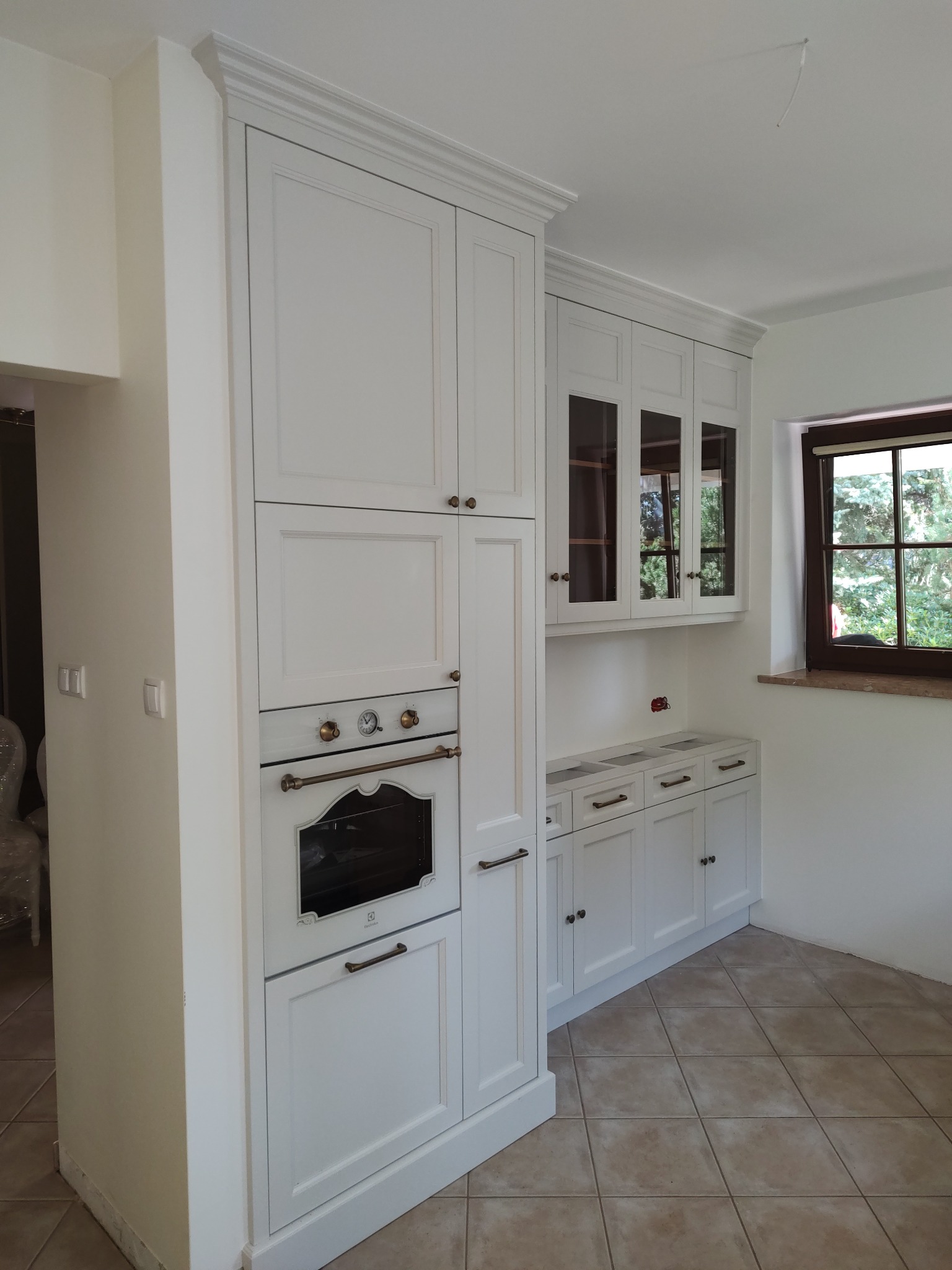 Klasyczna, biała zabudowa kuchenna z piekarnikiem Electrolux i przeszklonymi szafkami wiszącymi, widok z boku na tle okna i płytek podłogowych.