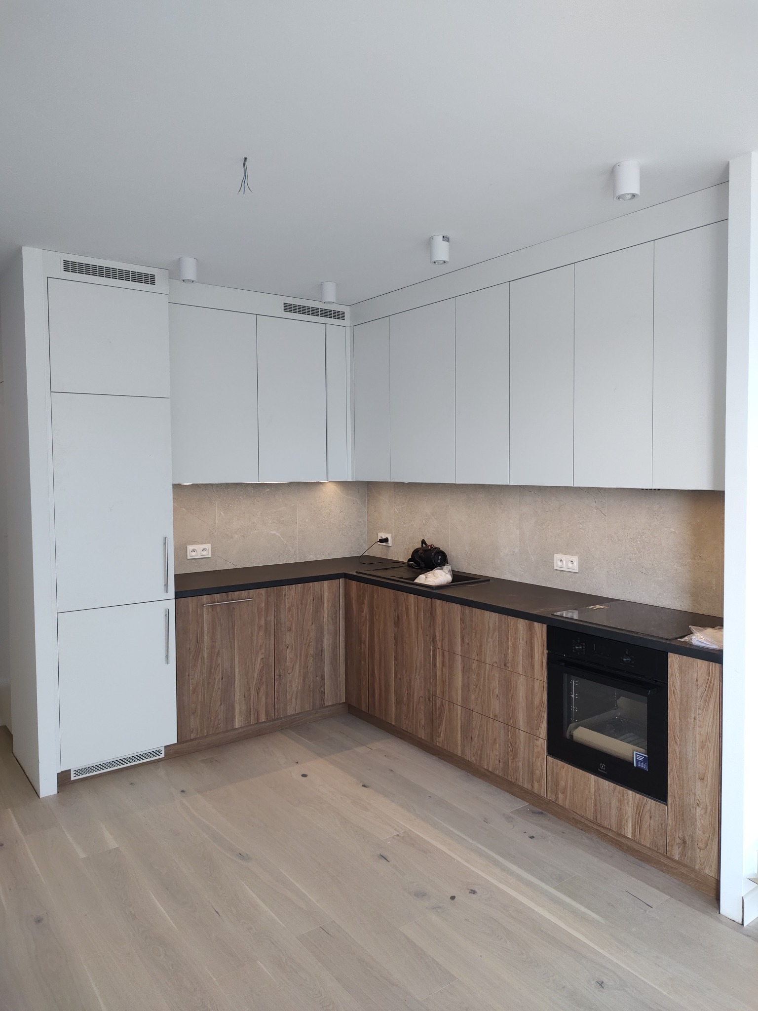 Nowoczesna kuchnia z białymi górnymi szafkami i drewnopodobnymi dolnymi szafkami, czarnym blatem, jasną podłogą, w tle marmurowa ściana i białe oświetlenie sufitowe.