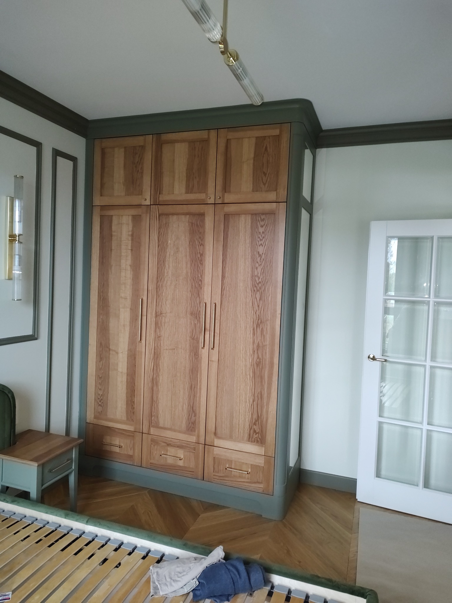 Drewniana szafa z trzema segmentami wbudowana w narożnik pokoju, wykończona w kolorze oliwkowym, z widocznymi detalami jak złote uchwyty i oświetlenie sufitowe.