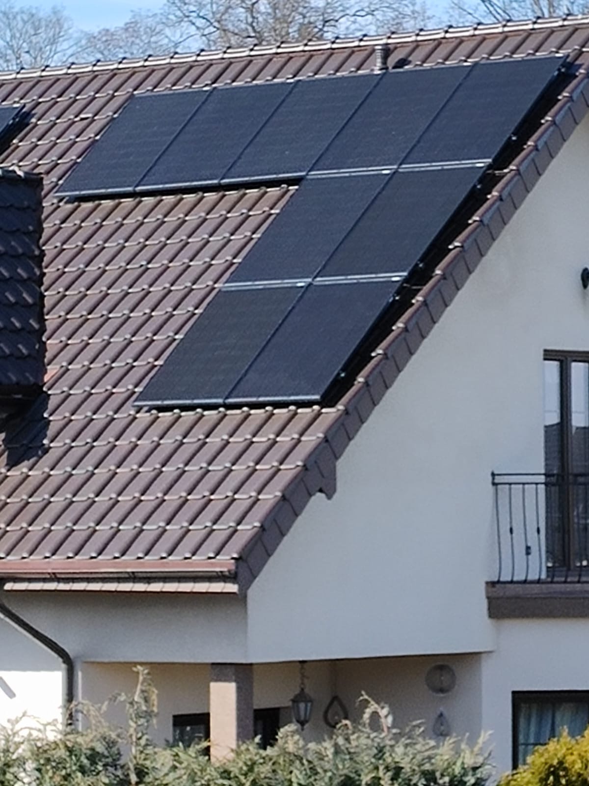 Panele słoneczne zamontowane na brązowym dachu z dachówki, widok na fragment elewacji budynku z balkonem.