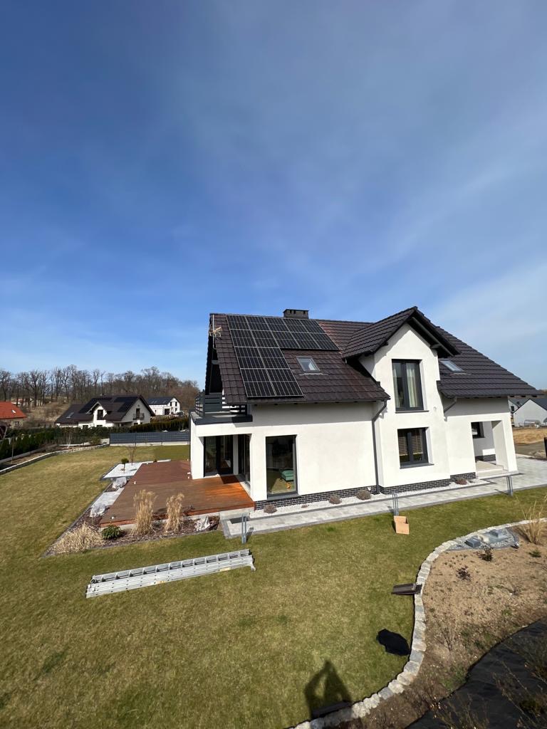 Nowoczesny dom jednorodzinny z brązowym dachem i panelami słonecznymi na części połaci, widoczny z perspektywy trawnika, na którym leży aluminiowa drabina.