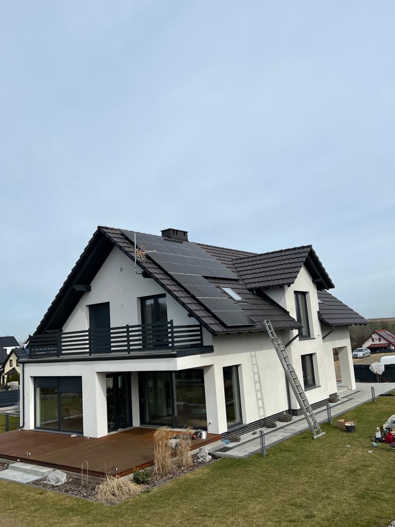 Nowoczesny dom jednorodzinny z brązowym dachem i panelami fotowoltaicznymi na dachu, drabina oparta o ścianę, widok z góry.