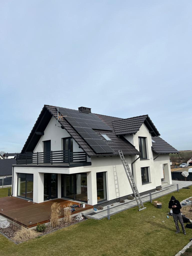 Nowoczesny dom jednorodzinny z panelami słonecznymi na dachu, drabina oparta o ścianę, widoczny pracownik w oddali, trawnik z elementami dekoracyjnymi.