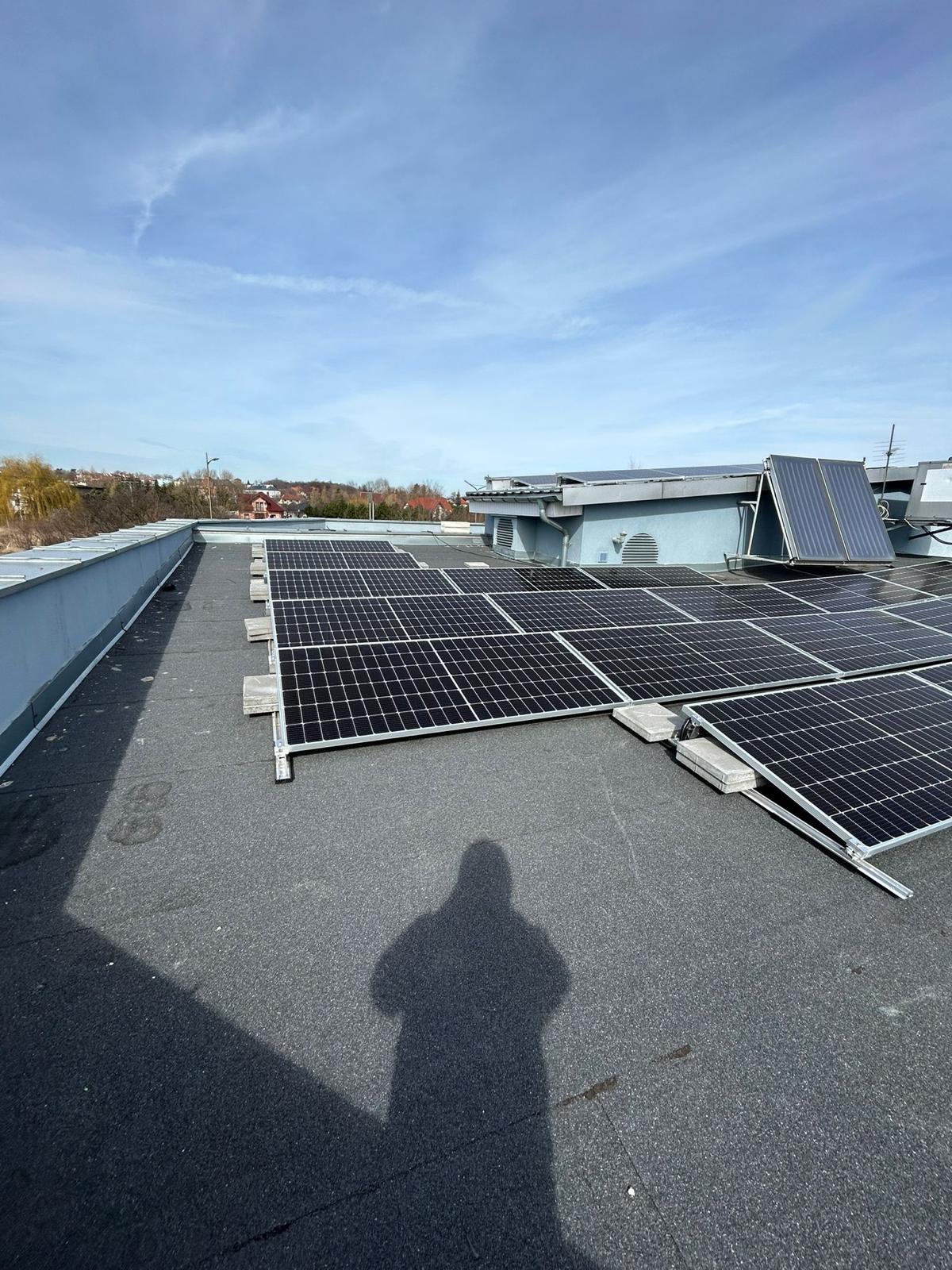 Panele słoneczne zamontowane na płaskim dachu budynku, widoczny cień osoby robiącej zdjęcie, w tle budynki i drzewa pod błękitnym niebem.