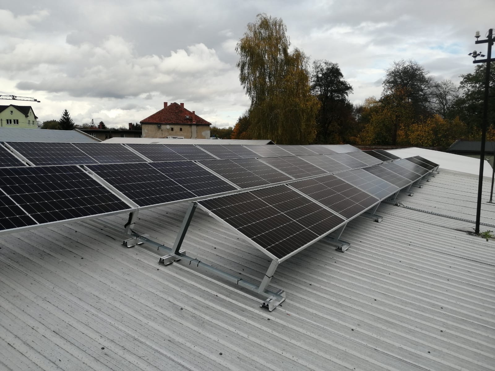 Rząd paneli słonecznych zamontowanych na metalowym dachu z widocznymi elementami mocującymi i budynkami w tle pod pochmurnym niebem.