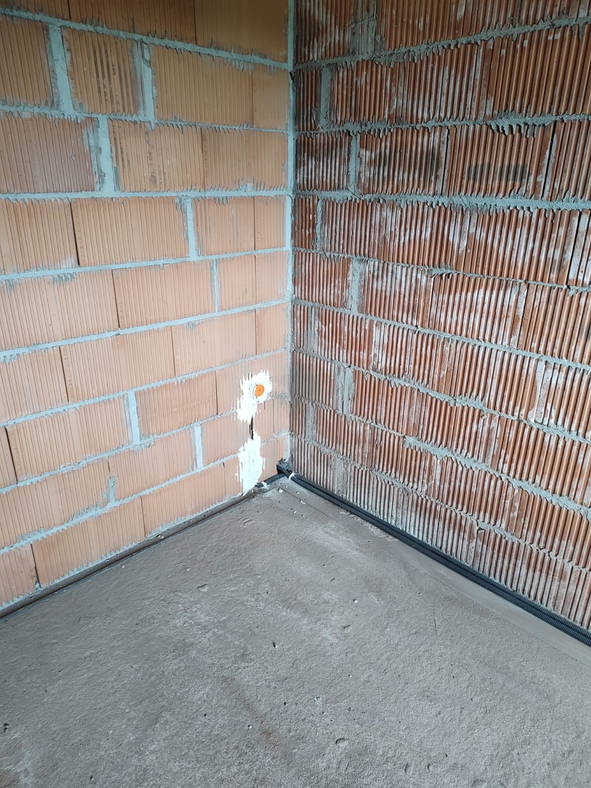 Surowy stan: Wewnętrzny róg pomieszczenia z murem z czerwonej cegły dziurawki, widoczna puszka instalacyjna i peszel przy podłodze z wylewką betonową.