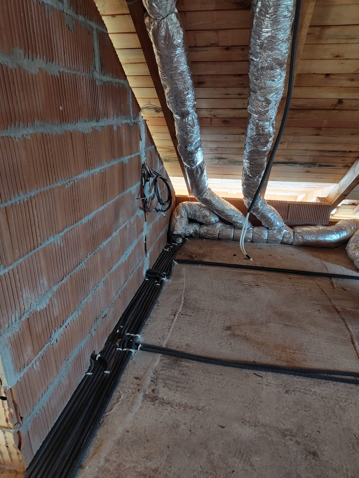 Instalacja przewodów w peszlach przy ceglanej ścianie na poddaszu w Karłowicach, widoczne także rury wentylacyjne pod drewnianym stropem.