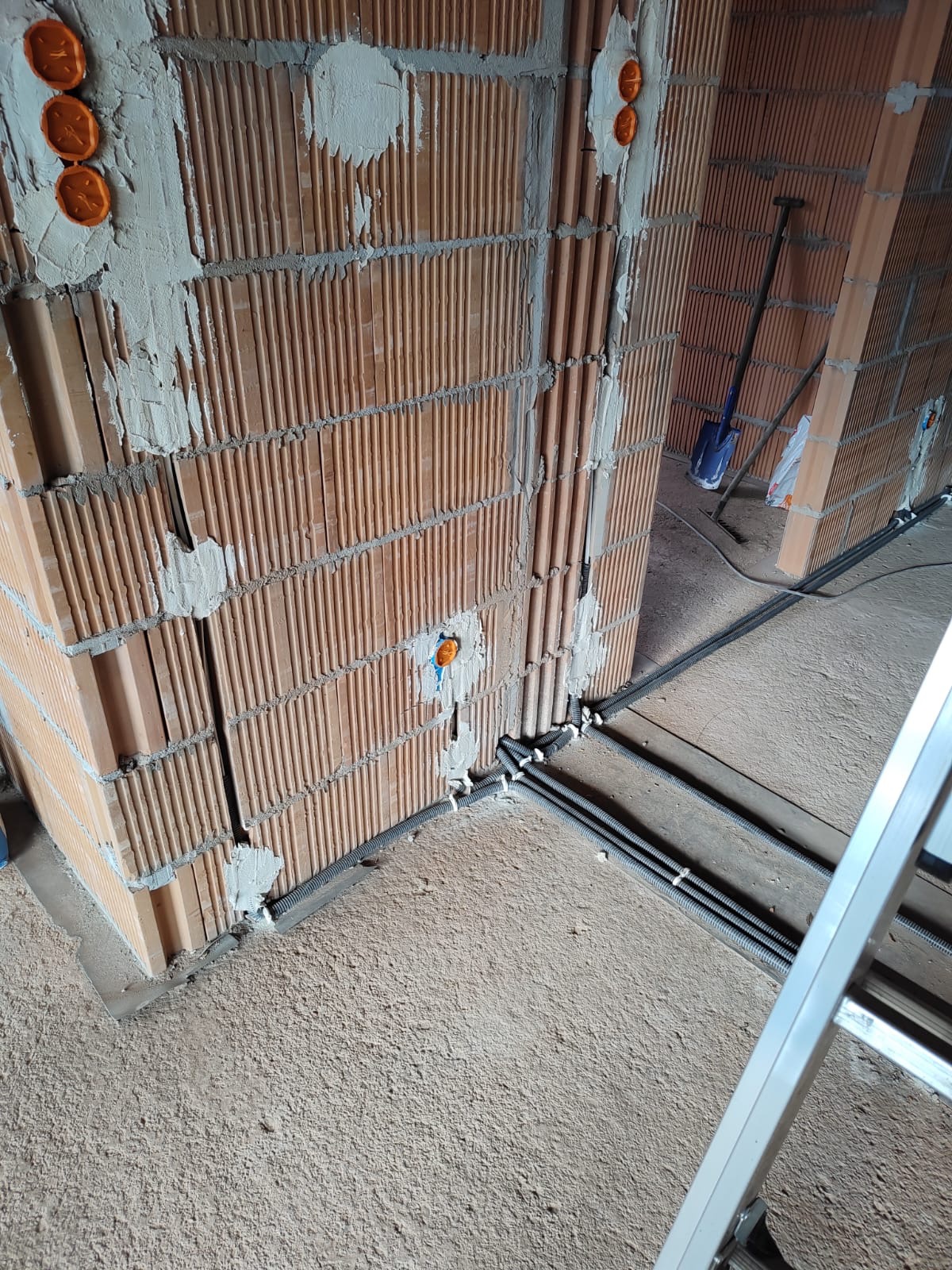 Wewnętrzne ściany z czerwonej cegły z widocznymi, częściowo zamurowanymi peszlami i puszkami instalacyjnymi, podłoga betonowa w surowym stanie, drabina aluminiowa w rogu kadru.