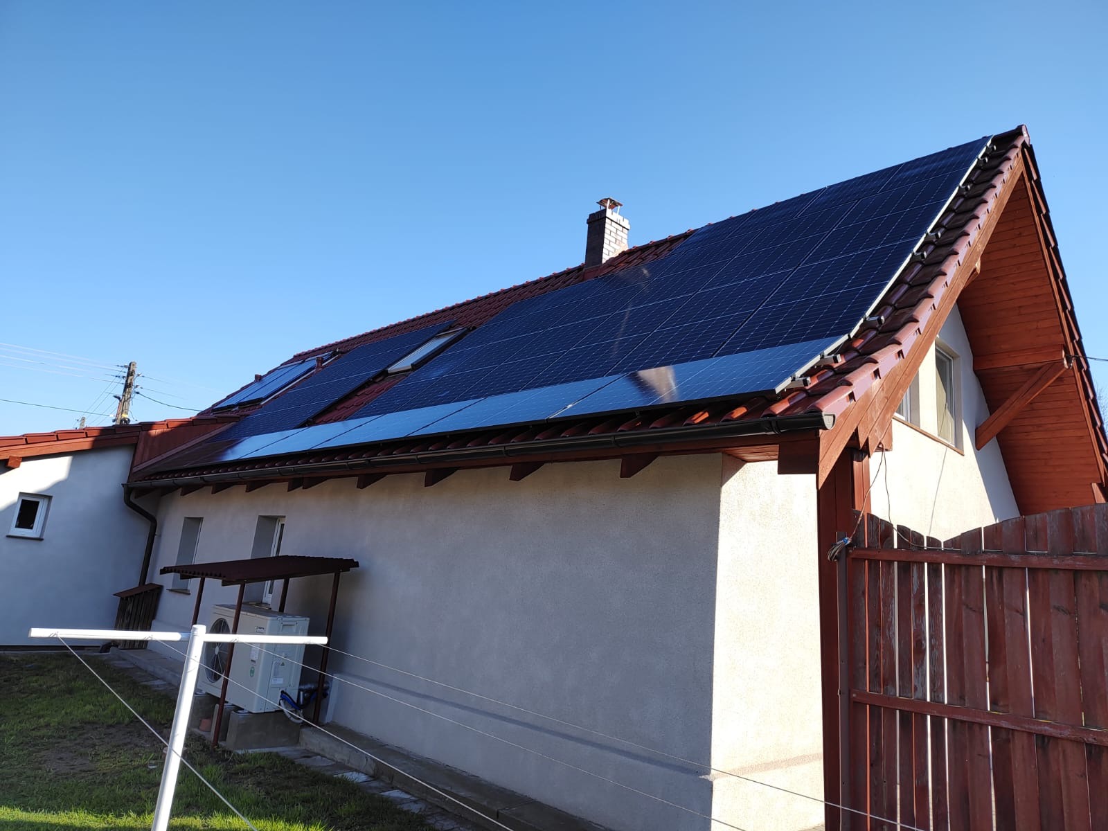 Dom jednorodzinny z panelami słonecznymi na dachu z czerwonej dachówki, widoczne okna dachowe i komin, na ścianie budynku zamontowana klimatyzacja.