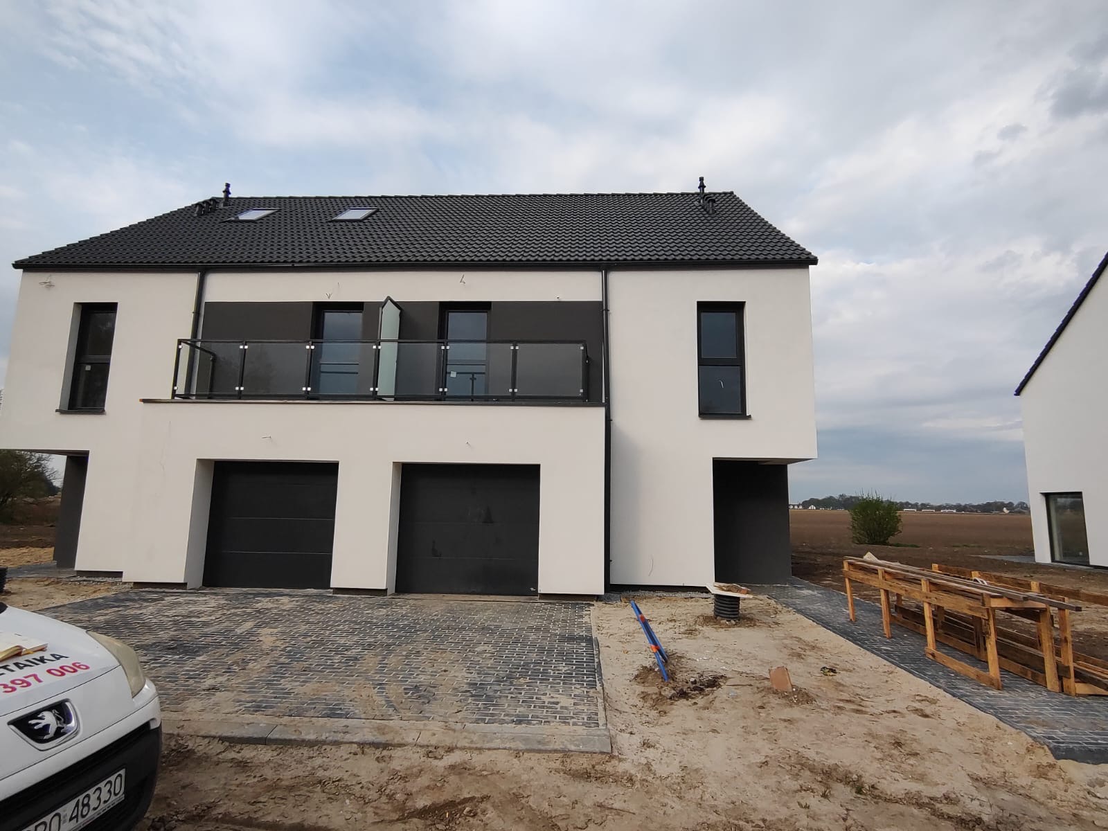 Nowoczesny, dwukondygnacyjny dom szeregowy z czarnymi bramami garażowymi i szklaną balustradą balkonu, kostka brukowa przed budynkiem w trakcie układania, widoczne elementy instalacji wystające...