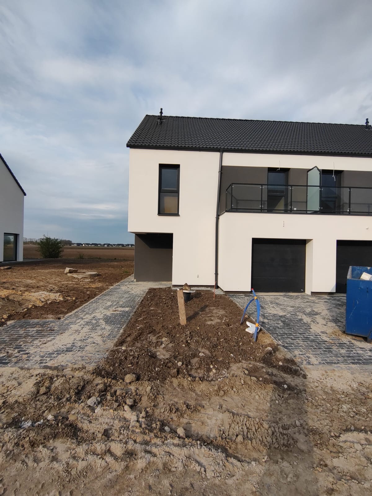 Nowoczesny szeregowiec z czarną stolarką okienną i balkonem z balustradą ze szkła, widok na świeżo ułożoną kostkę brukową i przygotowany teren pod trawnik, niebieski przewód na ziemi.