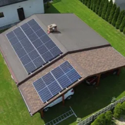 Widok z góry na dom z brązowym dachem pokrytym panelami słonecznymi; samochody zaparkowane pod zadaszeniem z boku domu; drabina oparta o ścianę zadaszenia.