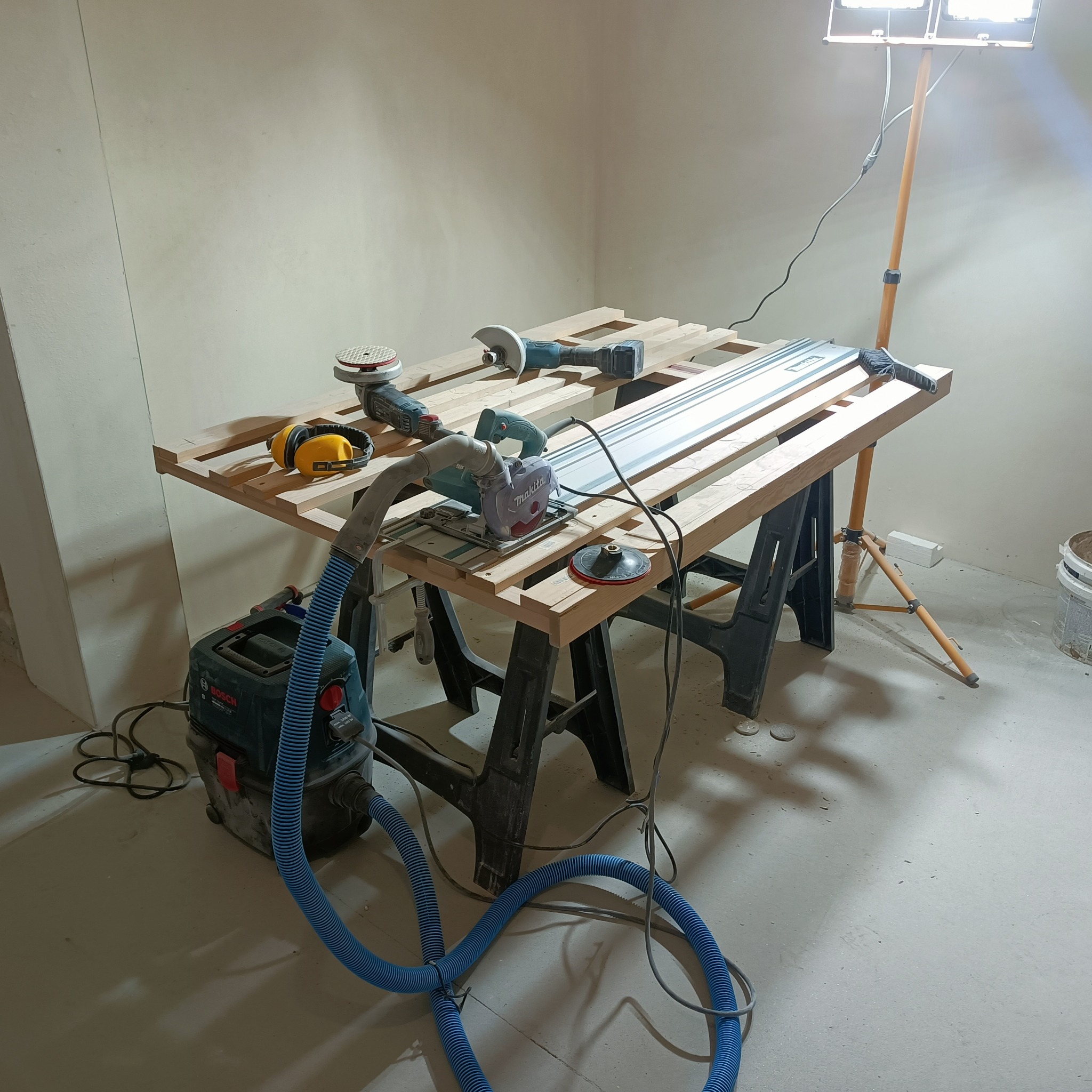 Stanowisko pracy z piłą tarczową, szlifierką i odkurzaczem przemysłowym Bosch, przygotowane do obróbki elementów drewnianych w surowym wnętrzu podczas remontu.