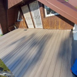 Pure NRG engineering - Nowo ułożony taras z desek kompozytowych w odcieniach szarości, z widoczną fakturą drewna, na tle drewnianego domu z oknami zabezpieczonymi kratami.