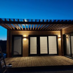 Pure NRG engineering - Pergola z drewnianą konstrukcją i szklanym dachem, oświetlona punktowymi lampami, w Bydgoszczy. W tle widoczne okna z roletami i elementy konstrukcyjne tarasu.