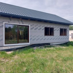 Pure NRG engineering - Dom w trakcie budowy, widoczne okna z brązowymi ramami zamontowane w ścianie pokrytej membraną ochronną, ciemna blachodachówka na dachu, w tle drzewa i niedokończona konstrukcja z białych bloczków.