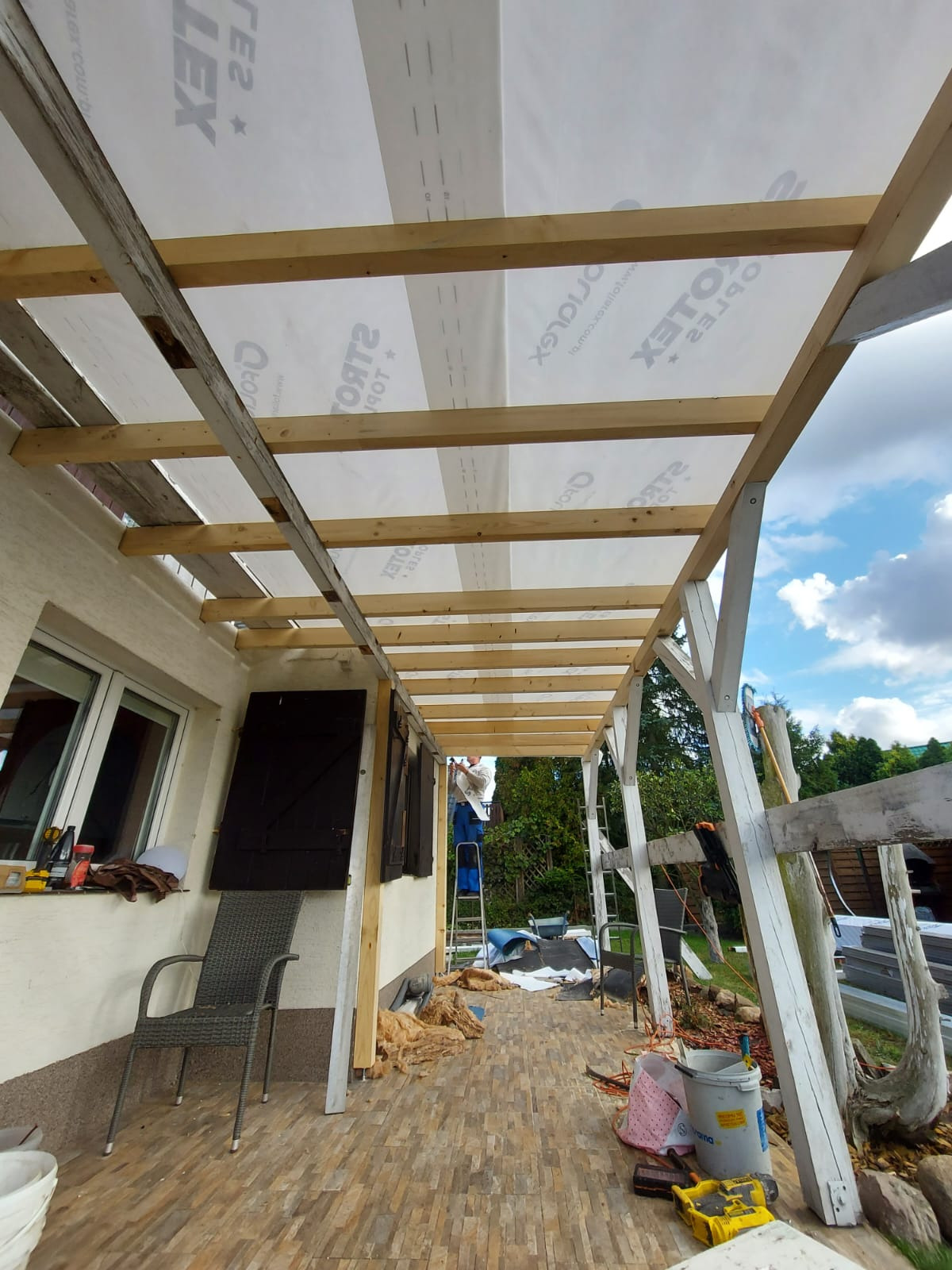 Montaż zadaszenia tarasu z drewnianą konstrukcją i folią izolacyjną, widoczny pracownik na drabinie, narzędzia oraz materiały budowlane na tarasie wyłożonym płytkami.