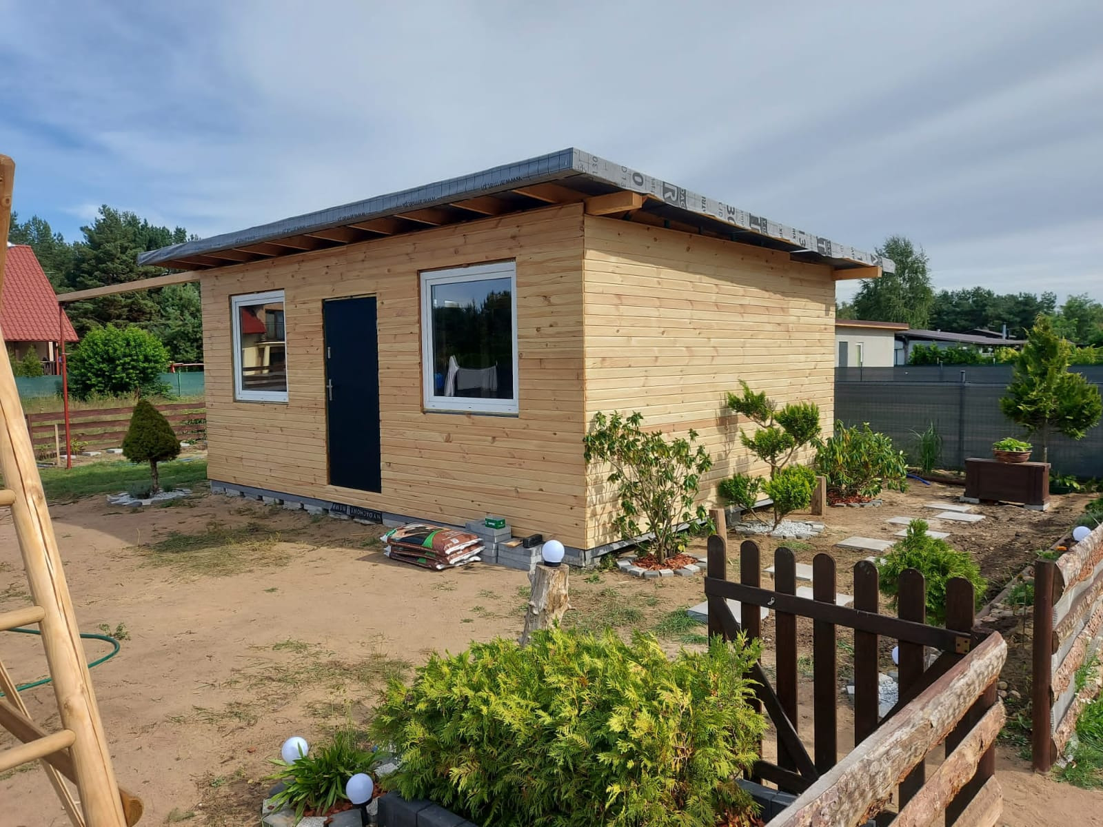 Nowo wybudowany drewniany domek z czarnymi drzwiami i białymi ramami okiennymi, widoczne worki z materiałami budowlanymi i drabina, całość otoczona ogrodem w trakcie aranżacji.