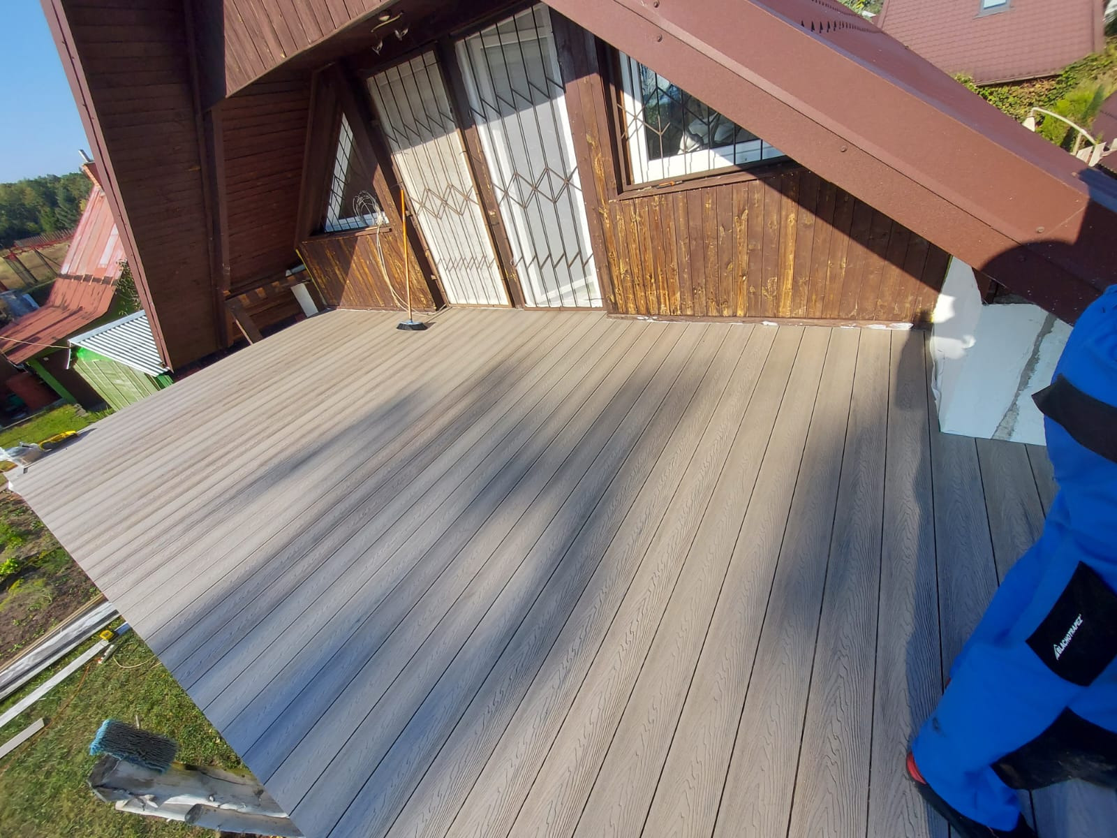 Nowo ułożony taras z desek kompozytowych w odcieniach szarości, z widoczną fakturą drewna, na tle drewnianego domu z oknami zabezpieczonymi kratami.