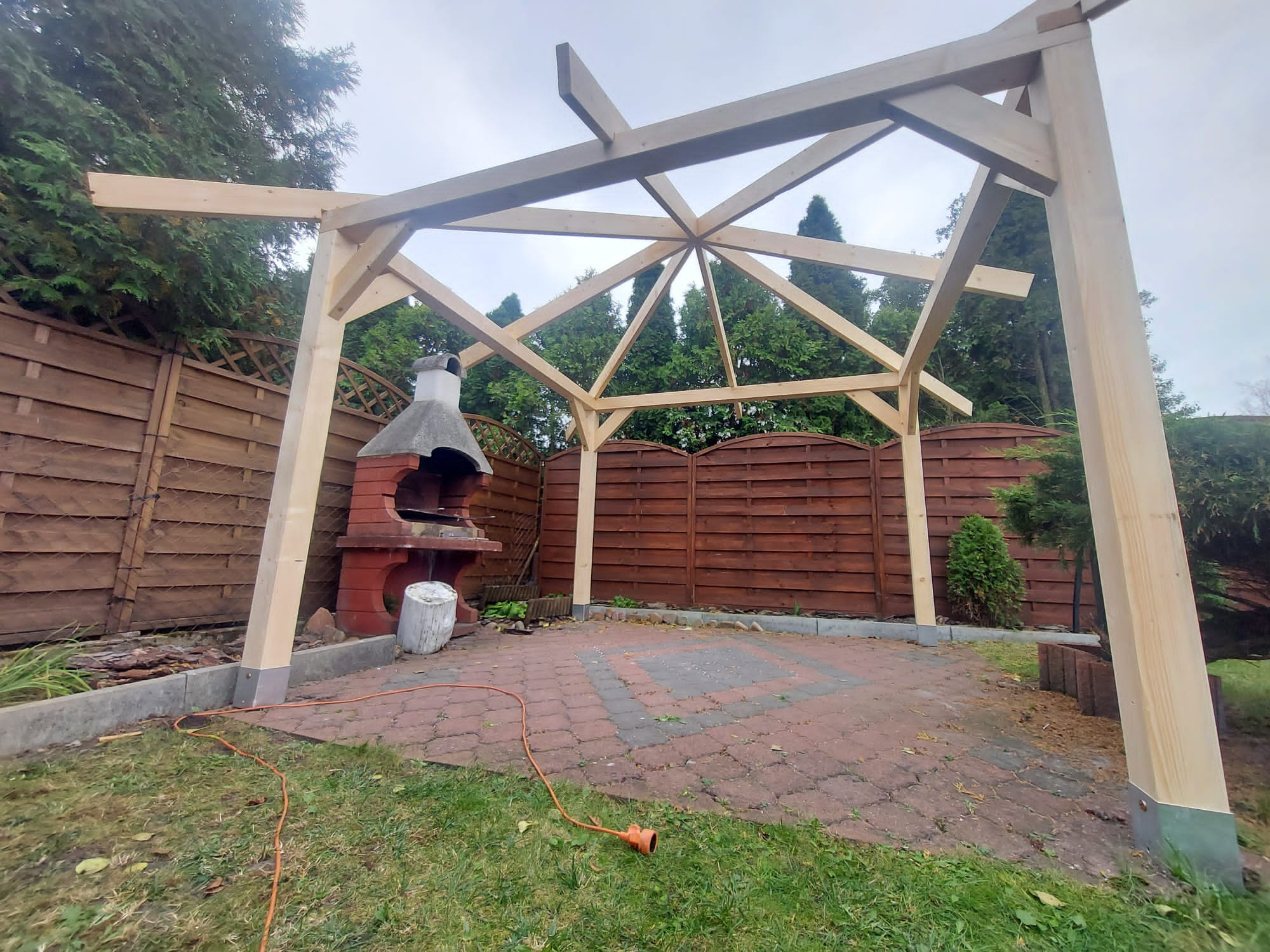 Konstrukcja drewnianej altany ogrodowej w trakcie budowy, widoczne elementy szkieletu dachu i słupy nośne, na tle ceglanego grilla i drewnianego ogrodzenia, z pomarańczowym przedłużaczem leżącym...