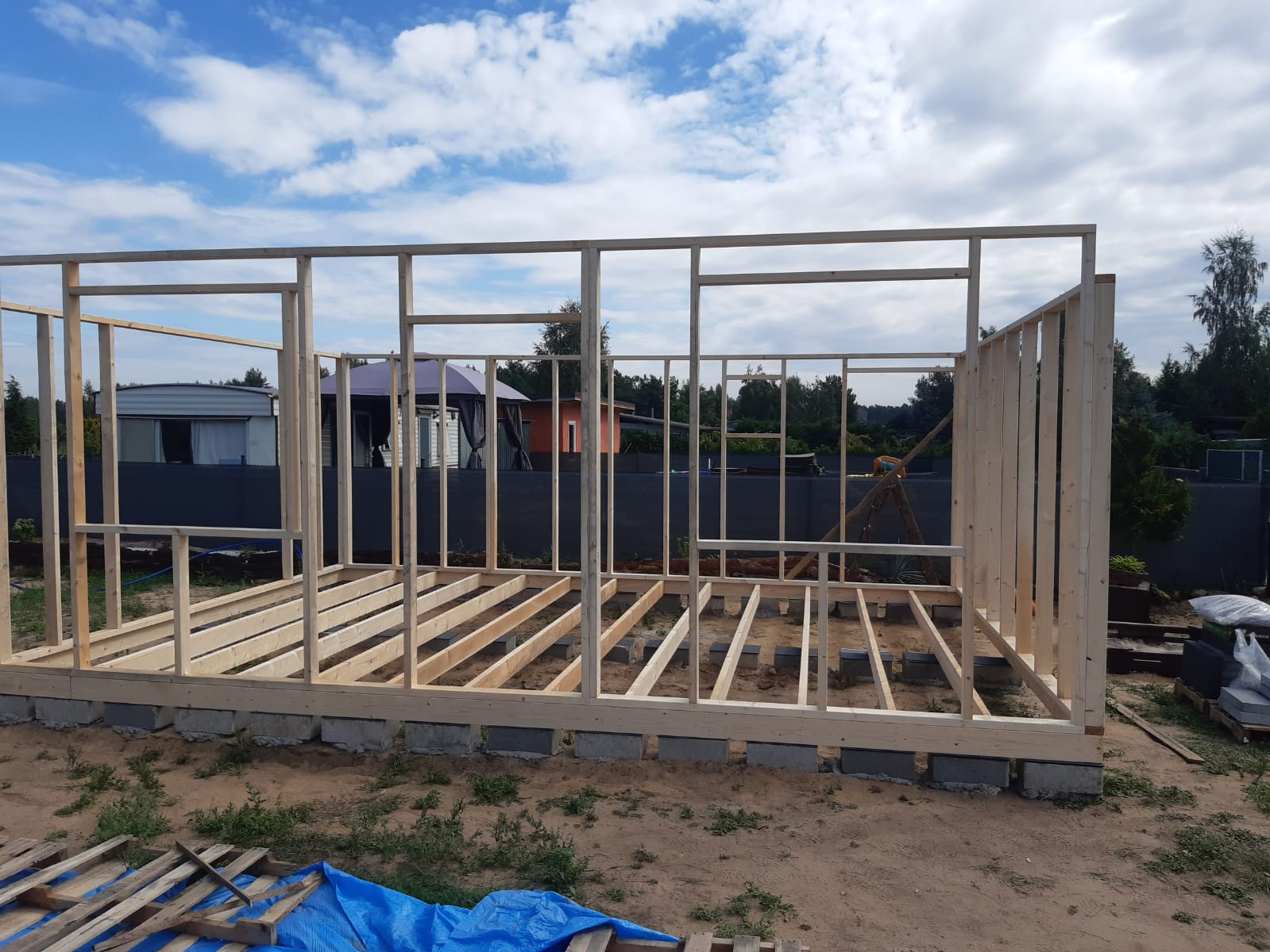 Konstrukcja szkieletowa budynku w trakcie budowy, widoczne drewniane belki i słupy na betonowej podmurówce, w tle zabudowa jednorodzinna i błękitne niebo z chmurami.