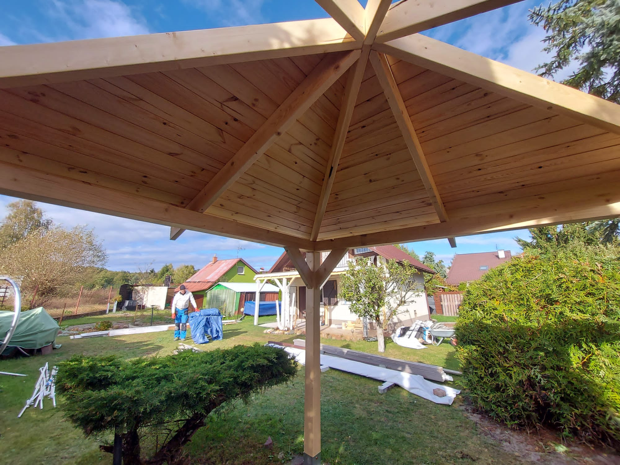 Widok od dołu na drewnianą konstrukcję zadaszenia w ogrodzie, z widocznym pracownikiem w tle i elementami przygotowanymi do dalszego montażu.