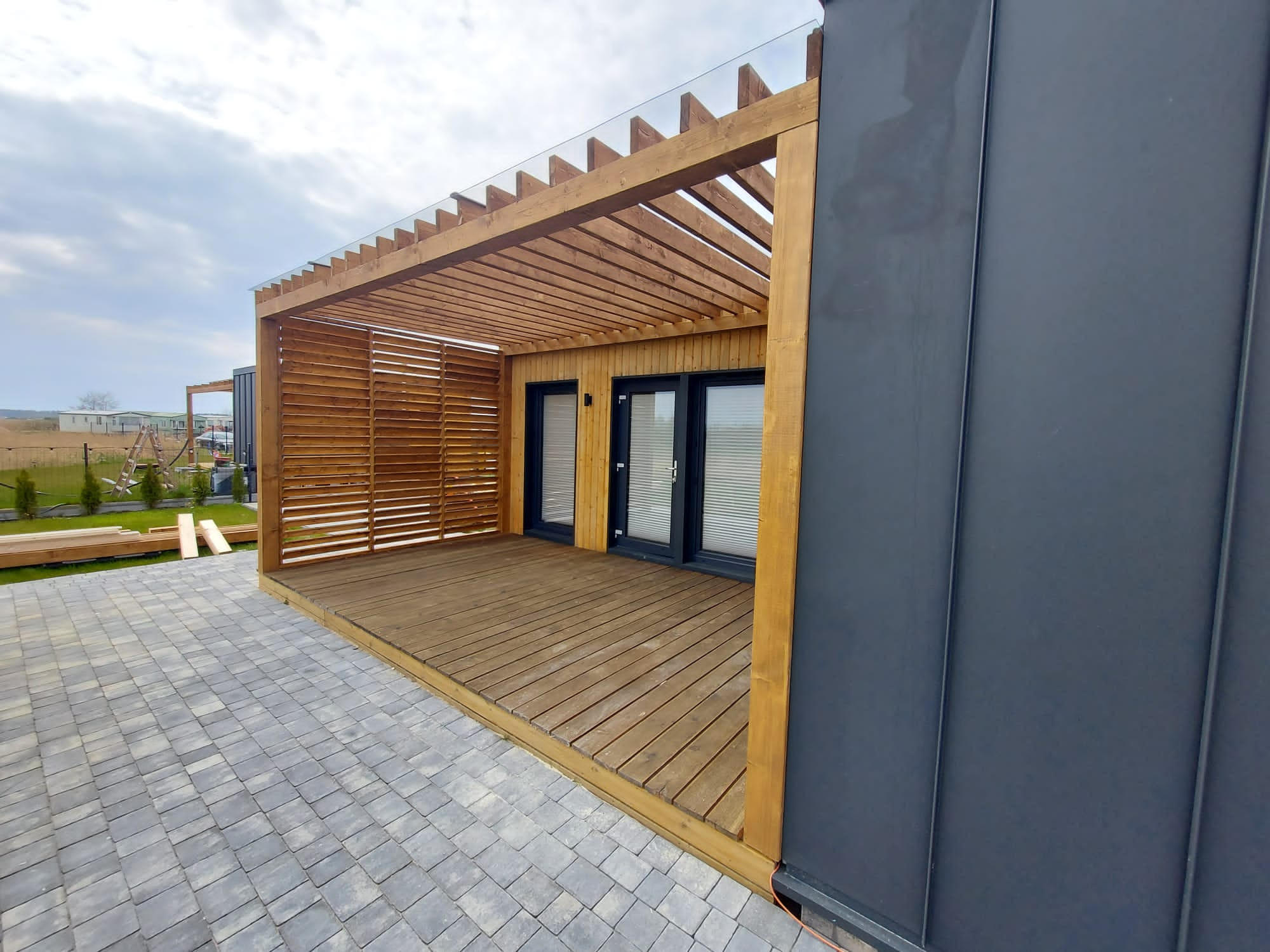 Nowoczesna drewniana pergola z zadaszeniem ze szkła na tarasie wyłożonym szarą kostką, widok z boku na szarą ścianę budynku.