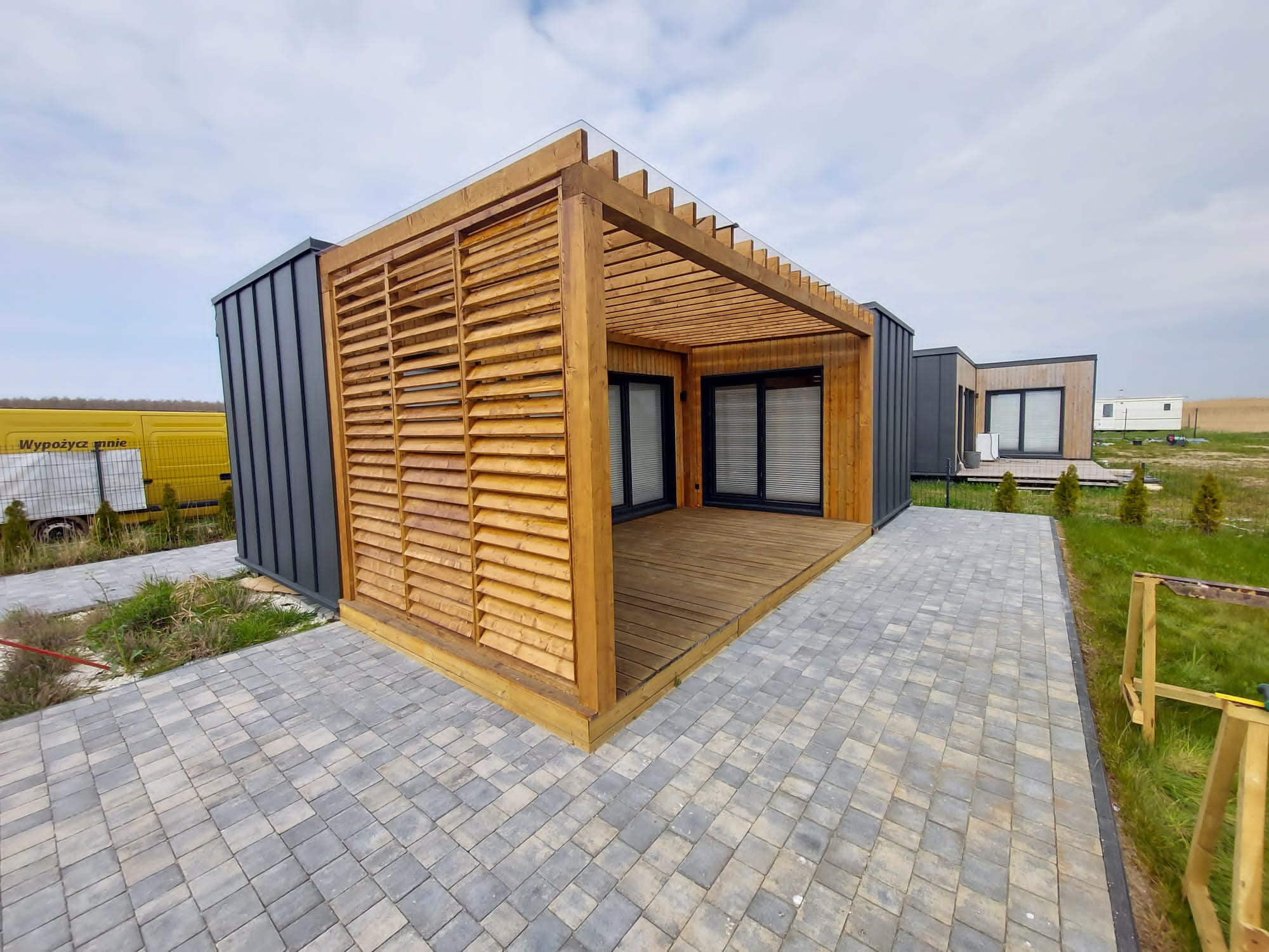 Nowoczesny modułowy domek z drewnianą pergolą i tarasem, szare panele elewacyjne, kostka brukowa na posesji, widok na okolicę.