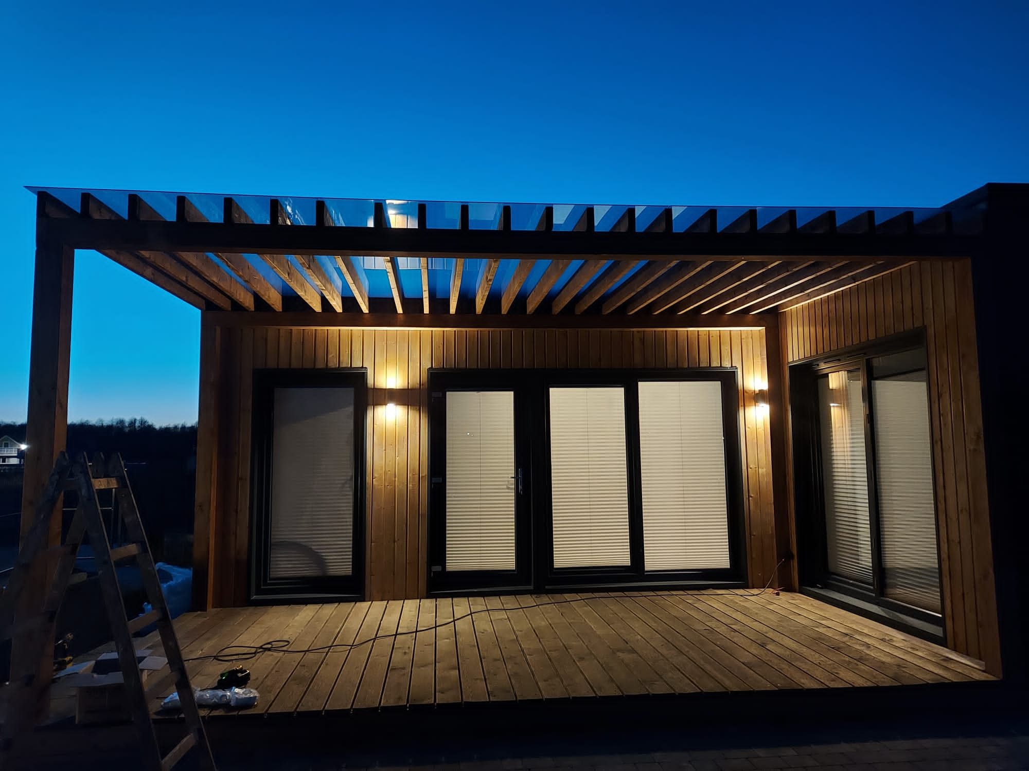 Pergola z drewnianą konstrukcją i szklanym dachem, oświetlona punktowymi lampami, w Bydgoszczy. W tle widoczne okna z roletami i elementy konstrukcyjne tarasu.