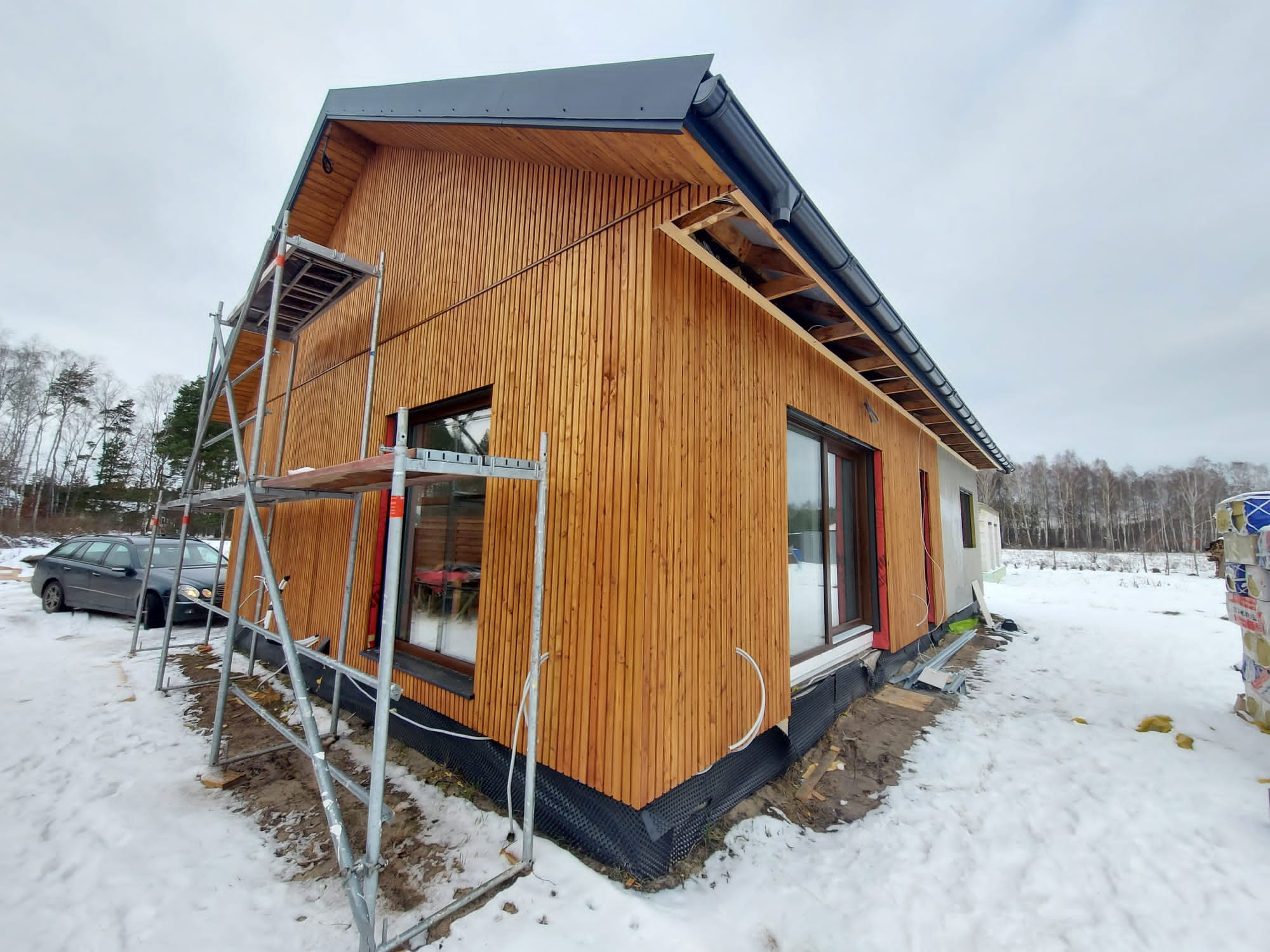 Dom w trakcie montażu drewnianej elewacji pionowej, widoczne rusztowanie, okna z ciemnymi ramami i fragment dachu z rynnami, teren pokryty śniegiem.