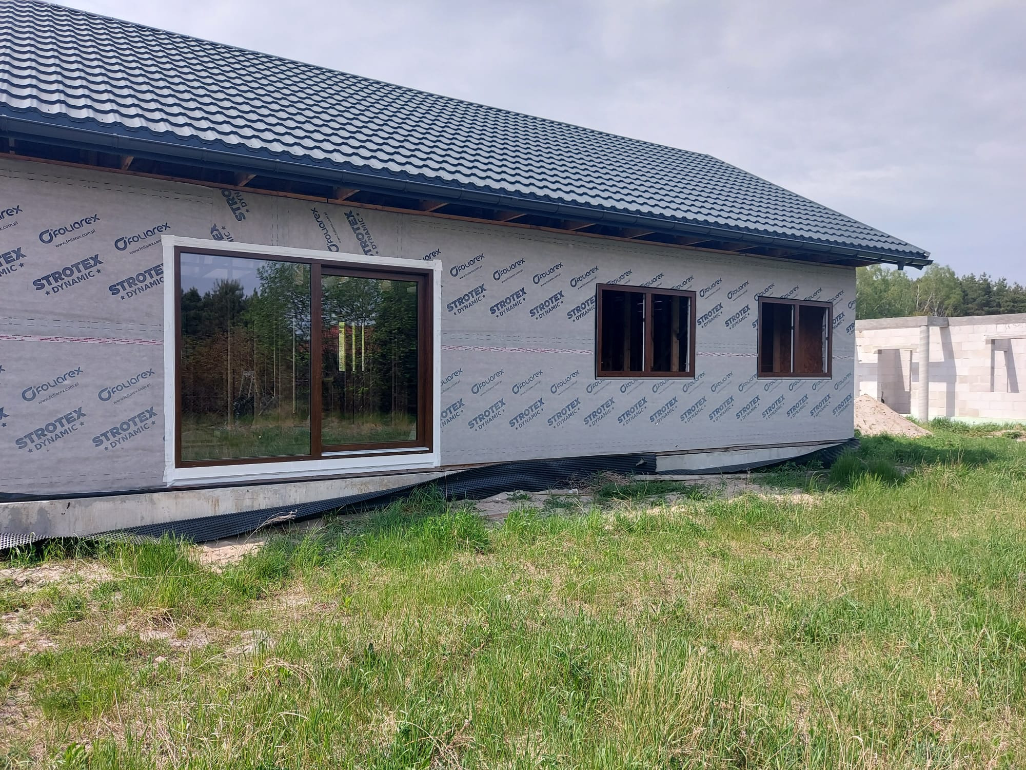 Dom w trakcie budowy, widoczne okna z brązowymi ramami zamontowane w ścianie pokrytej membraną ochronną, ciemna blachodachówka na dachu, w tle drzewa i niedokończona konstrukcja z białych bloczków.