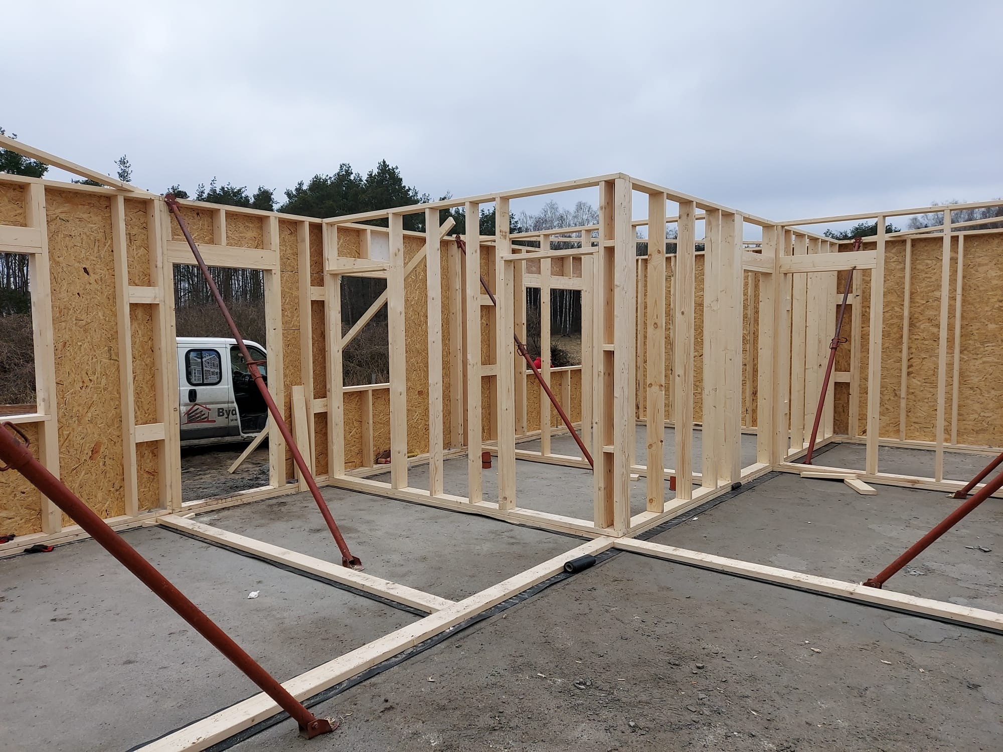 Konstrukcja szkieletowa domu w trakcie budowy, widoczne drewniane ramy ścian z płytami OSB, podparte czerwonymi stemplami budowlanymi, na surowym betonowym fundamencie, z widocznym samochodem...