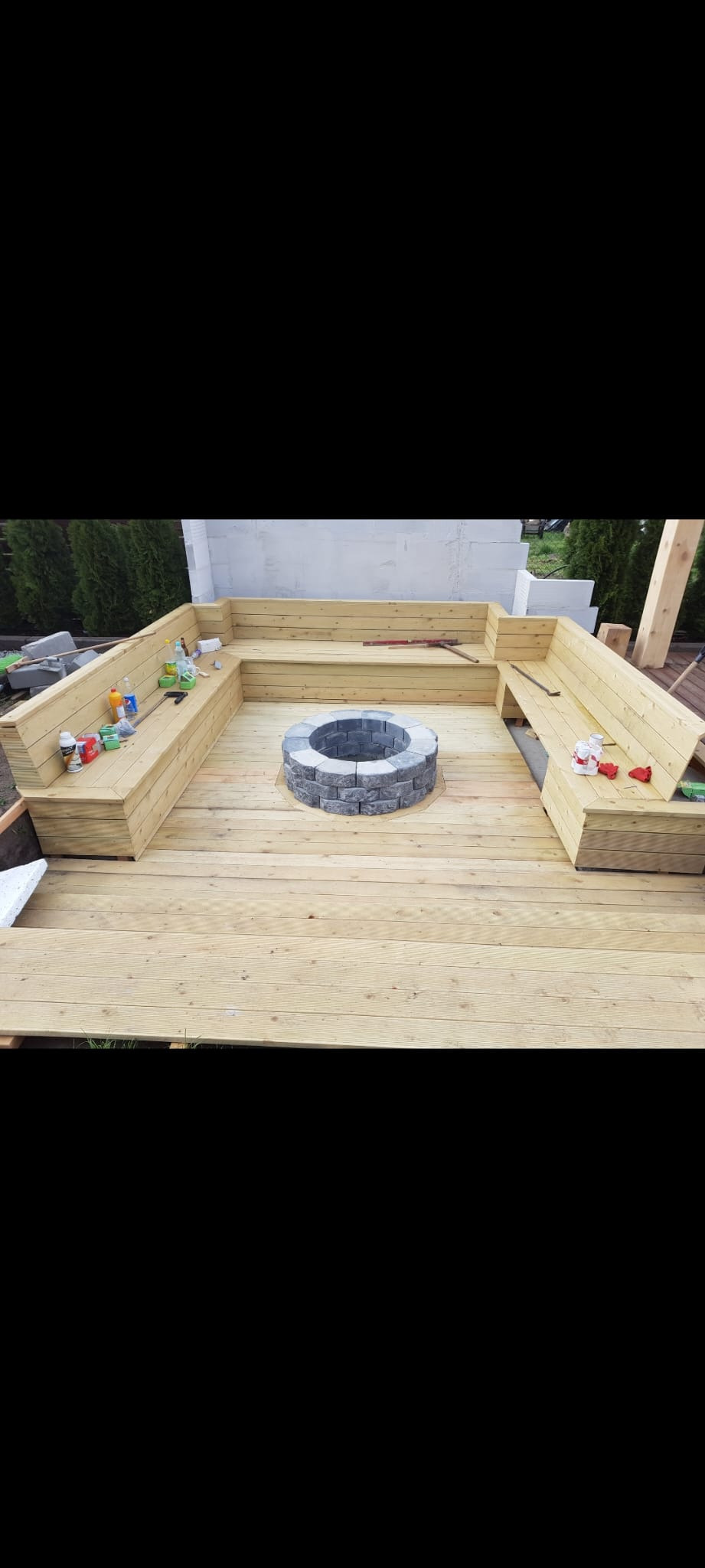 Drewniany taras z wbudowanymi ławkami i centralnym paleniskiem z szarej kostki, widoczne narzędzia i materiały budowlane