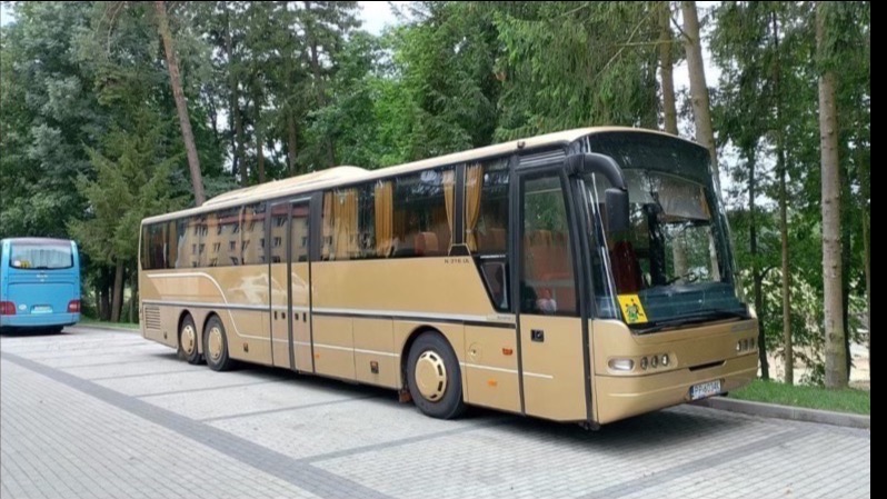 Złoty autokar turystyczny zaparkowany na parkingu z kostki brukowej, w tle zielone drzewa. Widoczny drugi, mniejszy, niebieski autobus.