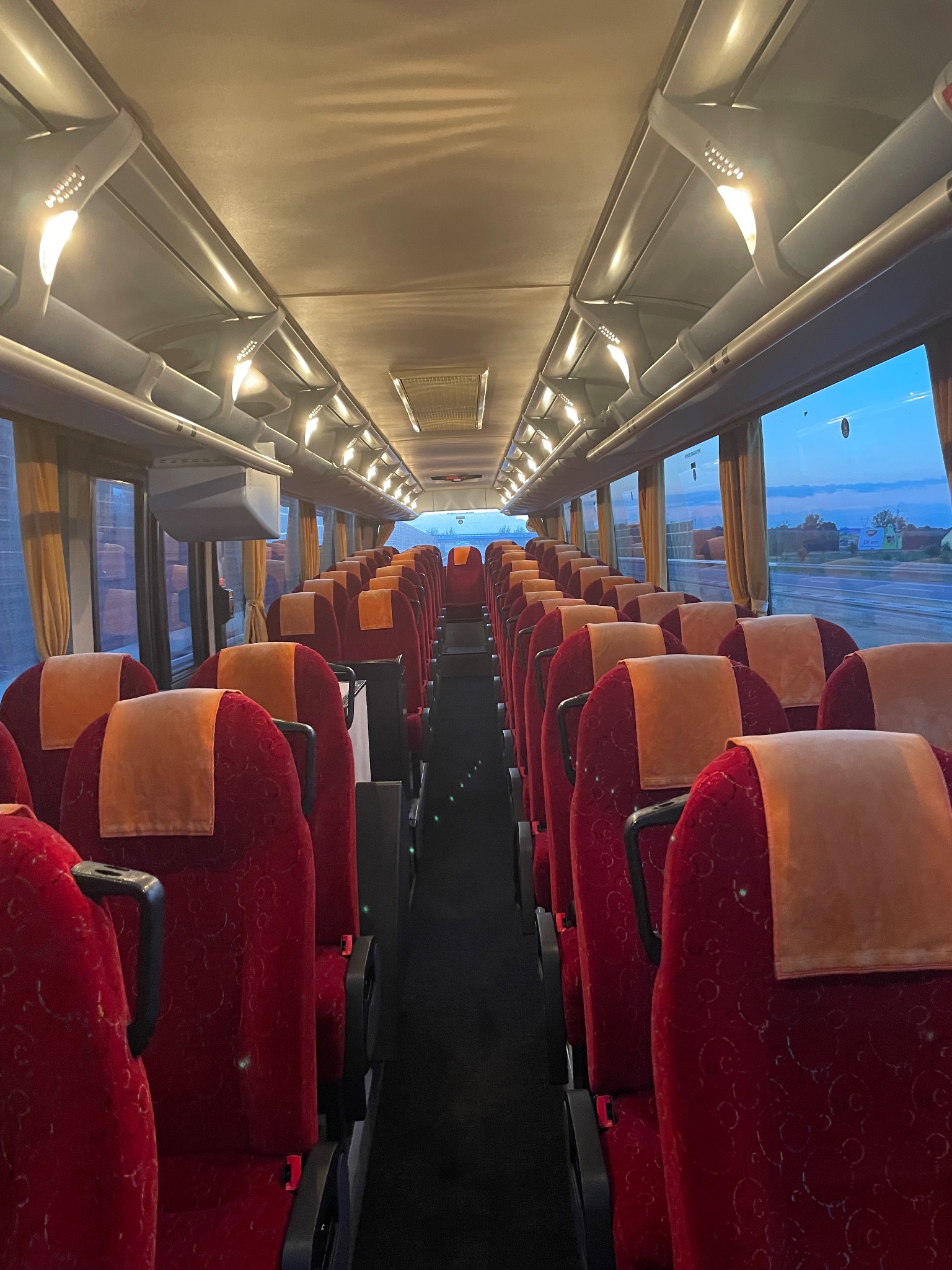 Wnętrze autobusu z rzędami czerwonych foteli z pomarańczowymi zagłówkami, widok na korytarz w kierunku okna z widokiem na krajobraz o zmierzchu.