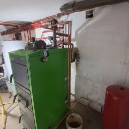 GoodHeat - Zielony piec SAS Varmo Plus z podłączeniami miedzianymi w kotłowni w Tarnobrzegu. Widoczne rury, zbiornik wyrównawczy i biały zasobnik na wodę.
