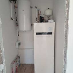 GoodHeat - Biała, kompaktowa pompa ciepła Panasonic Aquarea Smart Cloud w kotłowni z drewnopodobną podłogą, widoczne rury i zbiornik buforowy. Ściany w trakcie wykończenia.