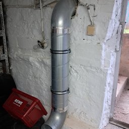 GoodHeat - Metalowa rura wentylacyjna z obejmami, zamontowana na nierównej, pomalowanej na biało ścianie w piwnicy. Widoczne instalacje elektryczne i czerwony koszyk REWE.