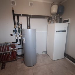 GoodHeat - Kompletna instalacja grzewcza z zbiornikiem i rozdzielaczem podłogówki, rury w izolacji, biała pompa ciepła w kotłowni na szarych płytkach podłogowych.