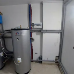 Dwa szare, pionowe zbiorniki na wodę z czerwonym i niebieskim zbiornikiem wyrównawczym u góry, widoczne rury instalacji, skrzynka elektryczna na ścianie, otwory w ścianie pod rurami.