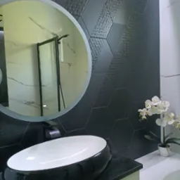 Nowoczesna łazienka z heksagonalnymi, grafitowymi płytkami z subtelnym wzorem, okrągłe lustro odbijające kabinę prysznicową, owalna umywalka nablatowa w kolorze czarno-białym i biała orchidea...