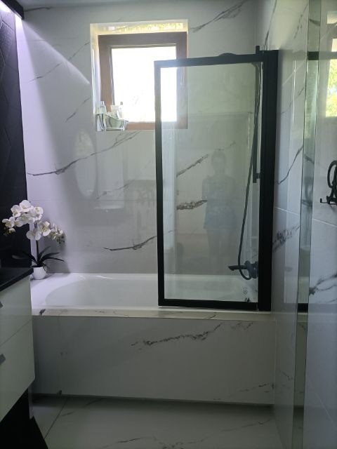 Nowoczesna łazienka z wanną obudowaną białymi płytkami imitującymi marmur, czarną ramą kabiny prysznicowej i białymi orchideami na brzegu wanny.