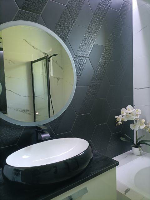 Nowoczesna łazienka z heksagonalnymi, grafitowymi płytkami z subtelnym wzorem, okrągłe lustro odbijające kabinę prysznicową, owalna umywalka nablatowa w kolorze czarno-białym i biała orchidea...