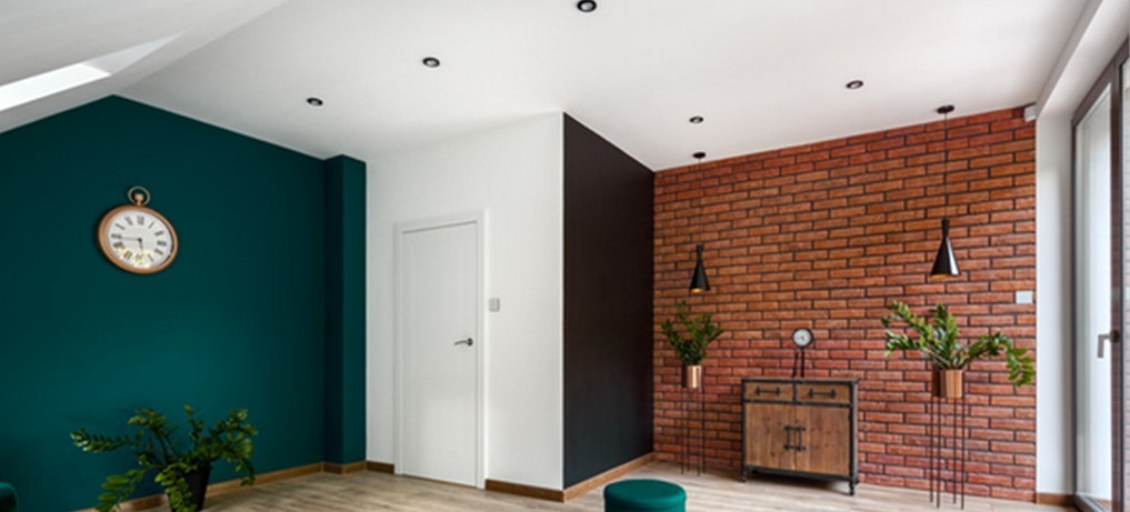 Nowoczesne wnętrze z kontrastującymi ścianami: butelkowa zieleń, biel, czerń i cegła. Zegar na ścianie, drewniana komoda, rośliny w doniczkach i punktowe oświetlenie sufitowe.