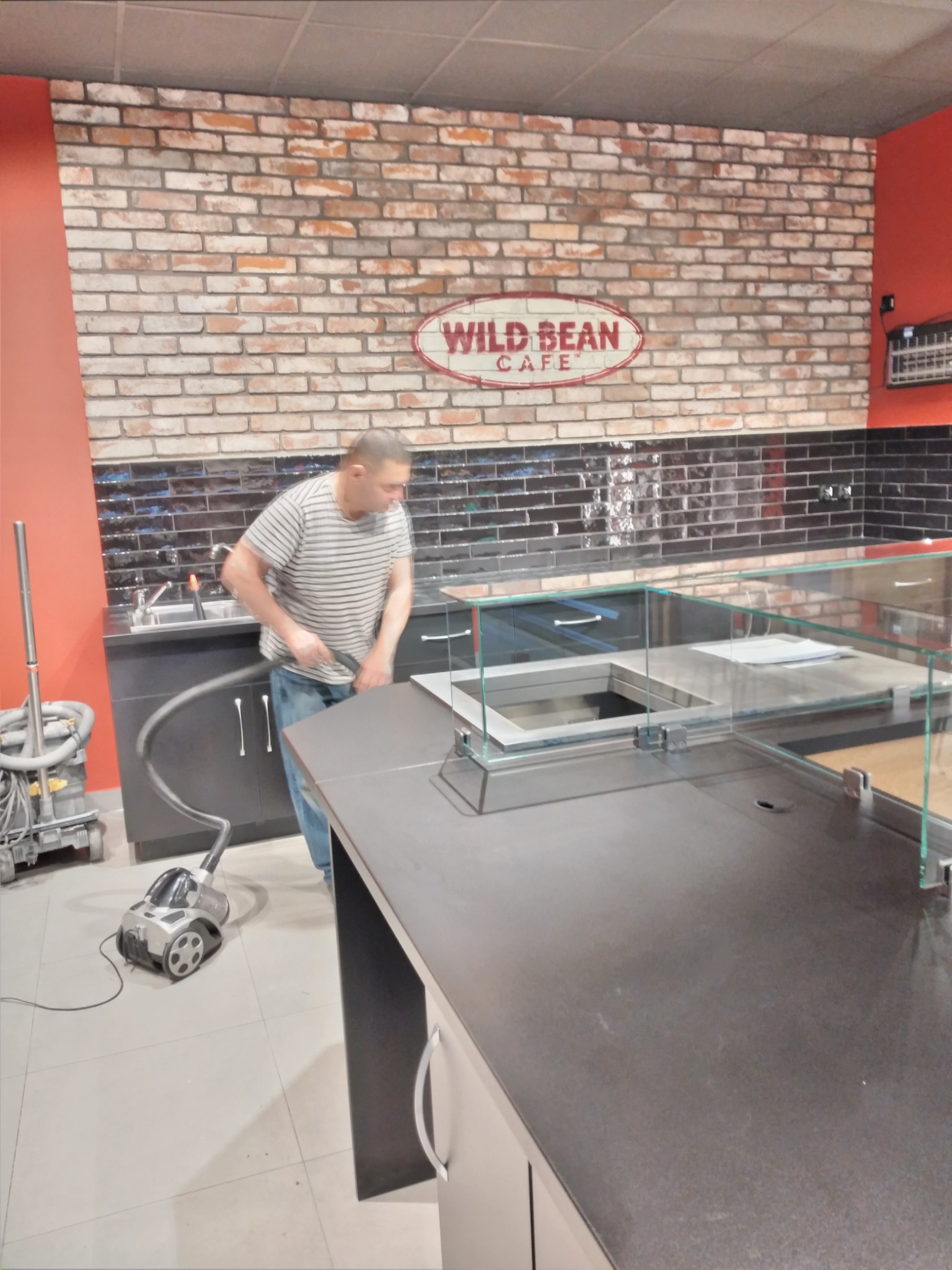 Mężczyzna odkurza wnętrze lokalu gastronomicznego, widoczny blat baru z szybą, ściana z cegieł i logo 'Wild Bean Cafe'.