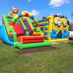 Kolorowy, dmuchany plac zabaw z motywem lwa i Minionków na trawiastym terenie, widoczny fragment budynku w tle.